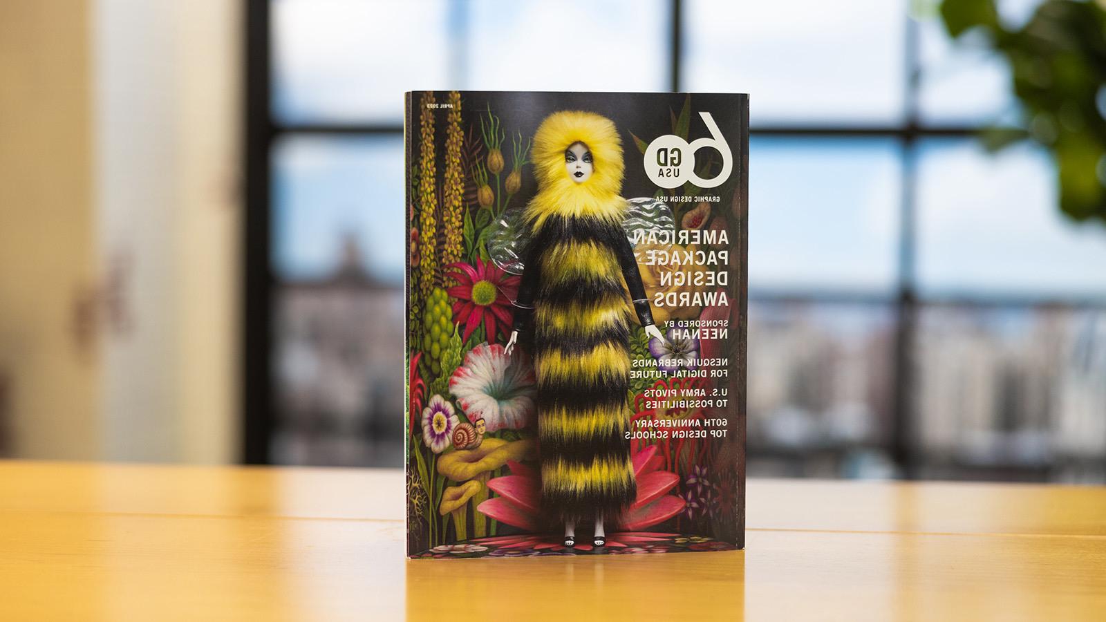 《美国平面设计》2023年4月刊笔直地放在木桌上. 封面上是一个女娃娃，穿着黑色和黄色的毛茸茸长裙，戴着兜帽. 她站在一个迷幻的花朵背景前.